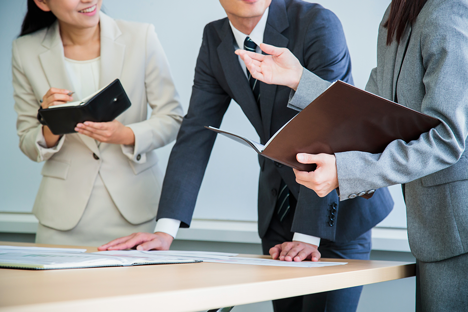 flow | 日本最初の品質マネジメントシステム認証機関 -日本化学キューエイ株式会社 Three professionals in business attire review documents on a table during a meeting.