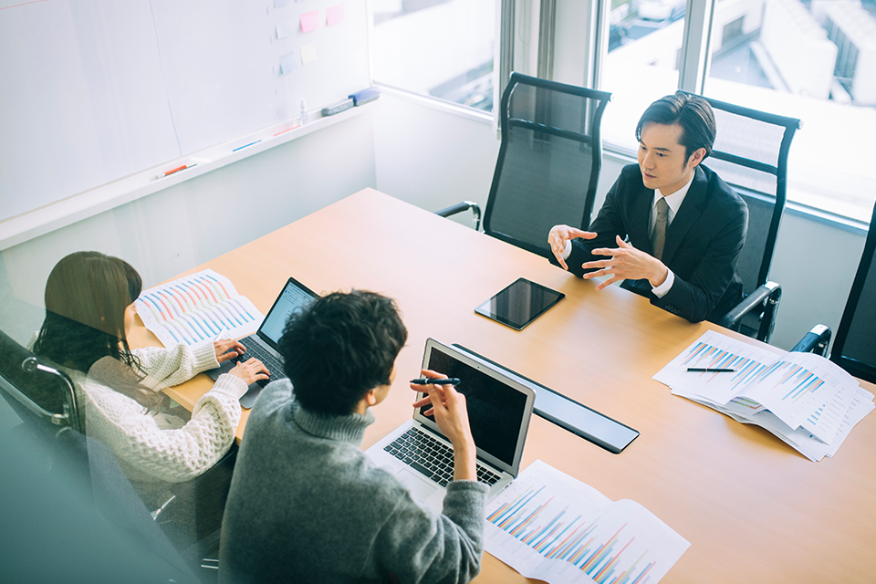 Three professionals in a meeting around a conference table with laptops and charts on the table.