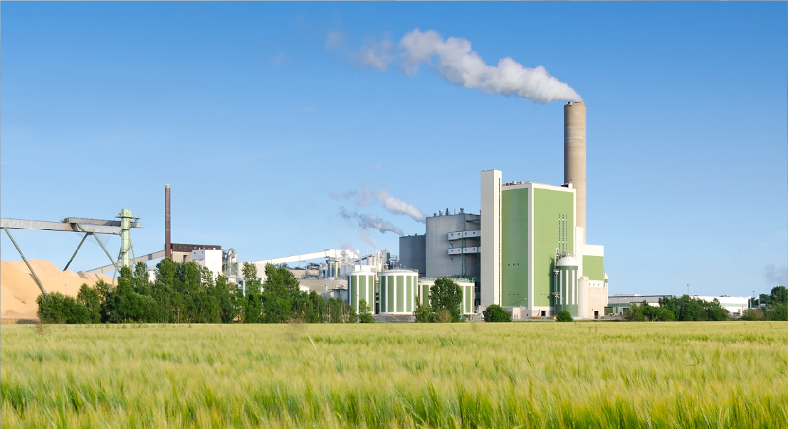 slide09 | 日本最初の品質マネジメントシステム認証機関 -日本化学キューエイ株式会社 Industrial power plant with a tall smokestack, green and gray buildings, and a line of trees under a blue sky.