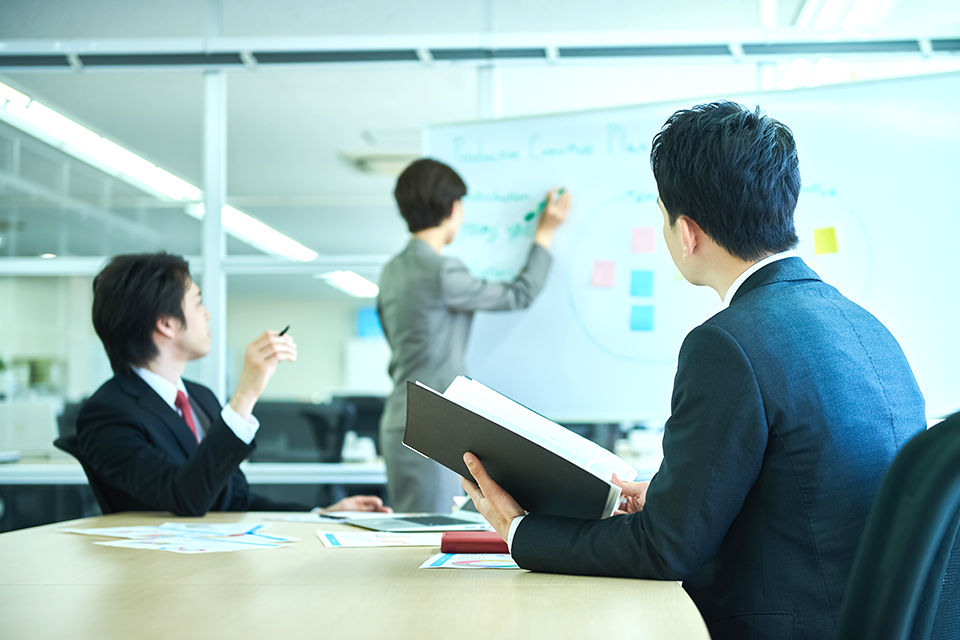 Three professionals in a modern office discuss a presentation, one at a whiteboard writing while two observe, one holding a folder.