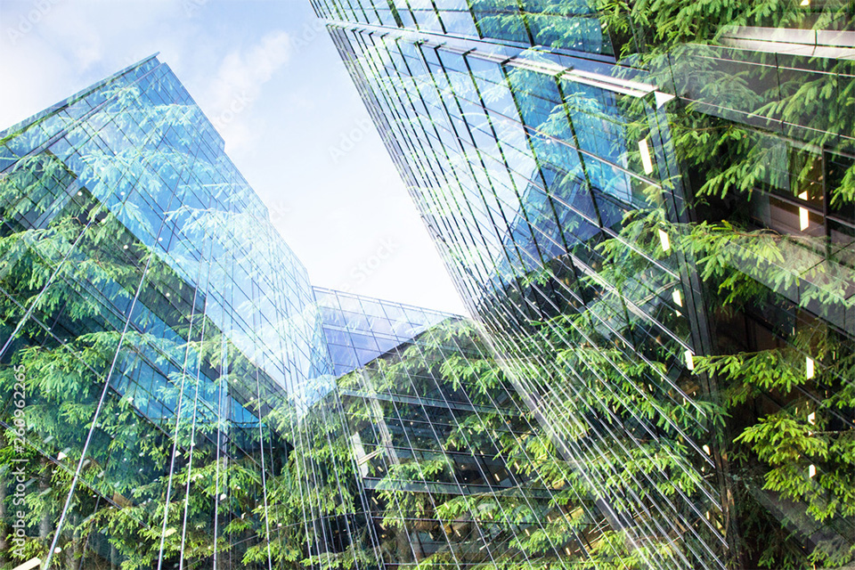 ghg01 | 日本最初の品質マネジメントシステム認証機関 -日本化学キューエイ株式会社 Low-angle view of glass office buildings with green trees reflected in the windows and a blue sky above.