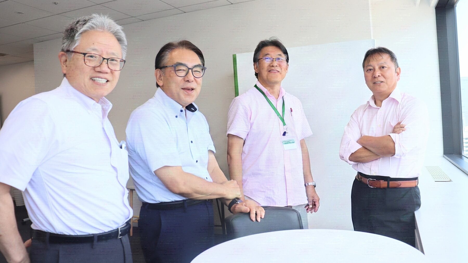 審査員1 | 日本最初の品質マネジメントシステム認証機関 -日本化学キューエイ株式会社 Four men in business casual attire stand together in an office near a round table, posing for a group photo and smiling at the camera.
