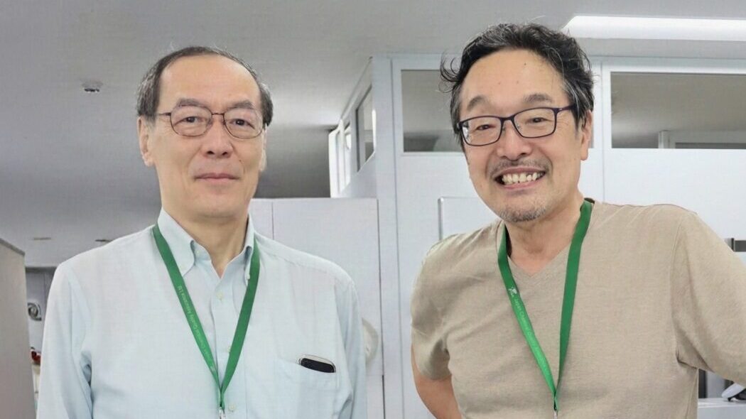 審査員2 | 日本最初の品質マネジメントシステム認証機関 -日本化学キューエイ株式会社 Two men with green lanyards and ID badges pose together in an office setting, smiling at the camera.