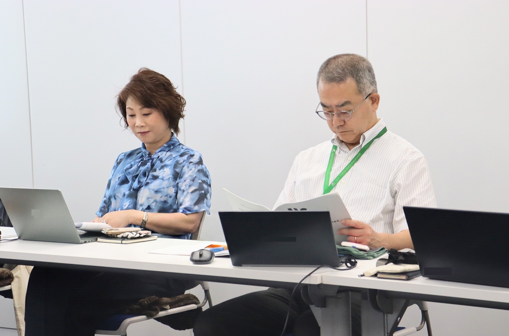 審査風景1 | 日本最初の品質マネジメントシステム認証機関 -日本化学キューエイ株式会社 Two professionals sit at a table with laptops and papers; woman on the left in a blue blouse and the man on the right with a green lanyard reads a document.