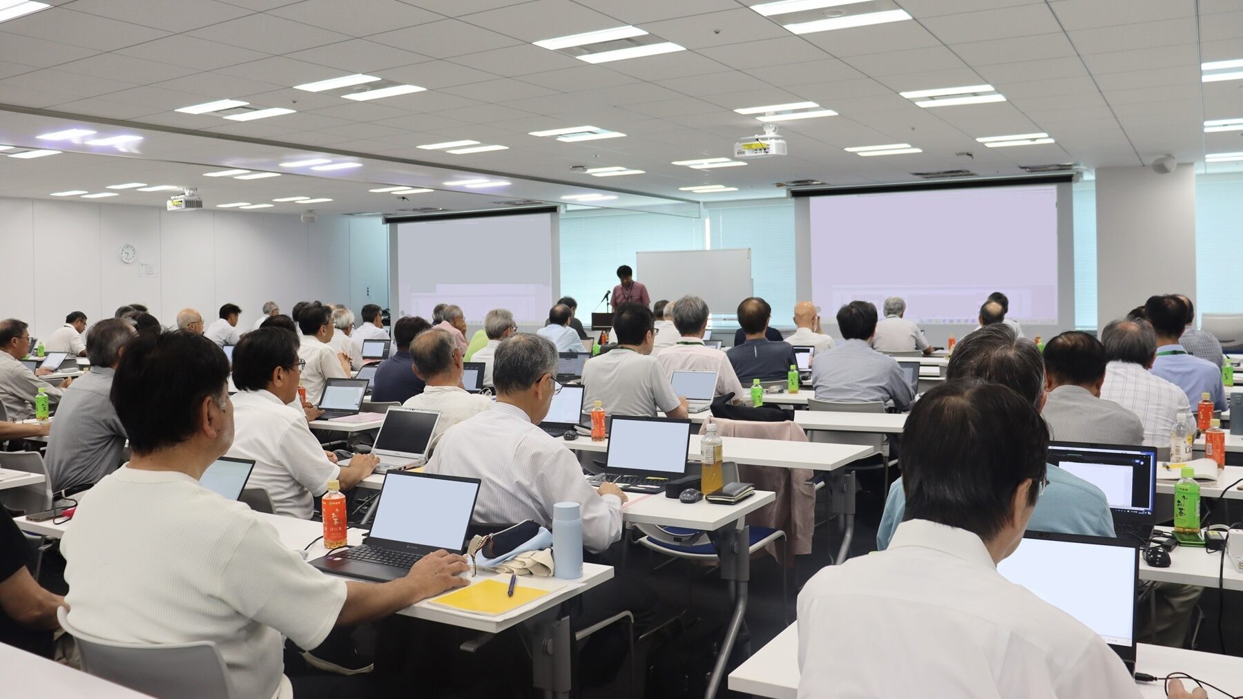 集合研修会 | 日本最初の品質マネジメントシステム認証機関 -日本化学キューエイ株式会社 Conference room full of attendees with laptops, a speaker at a podium near two projection screens at the front of the room, presenting.