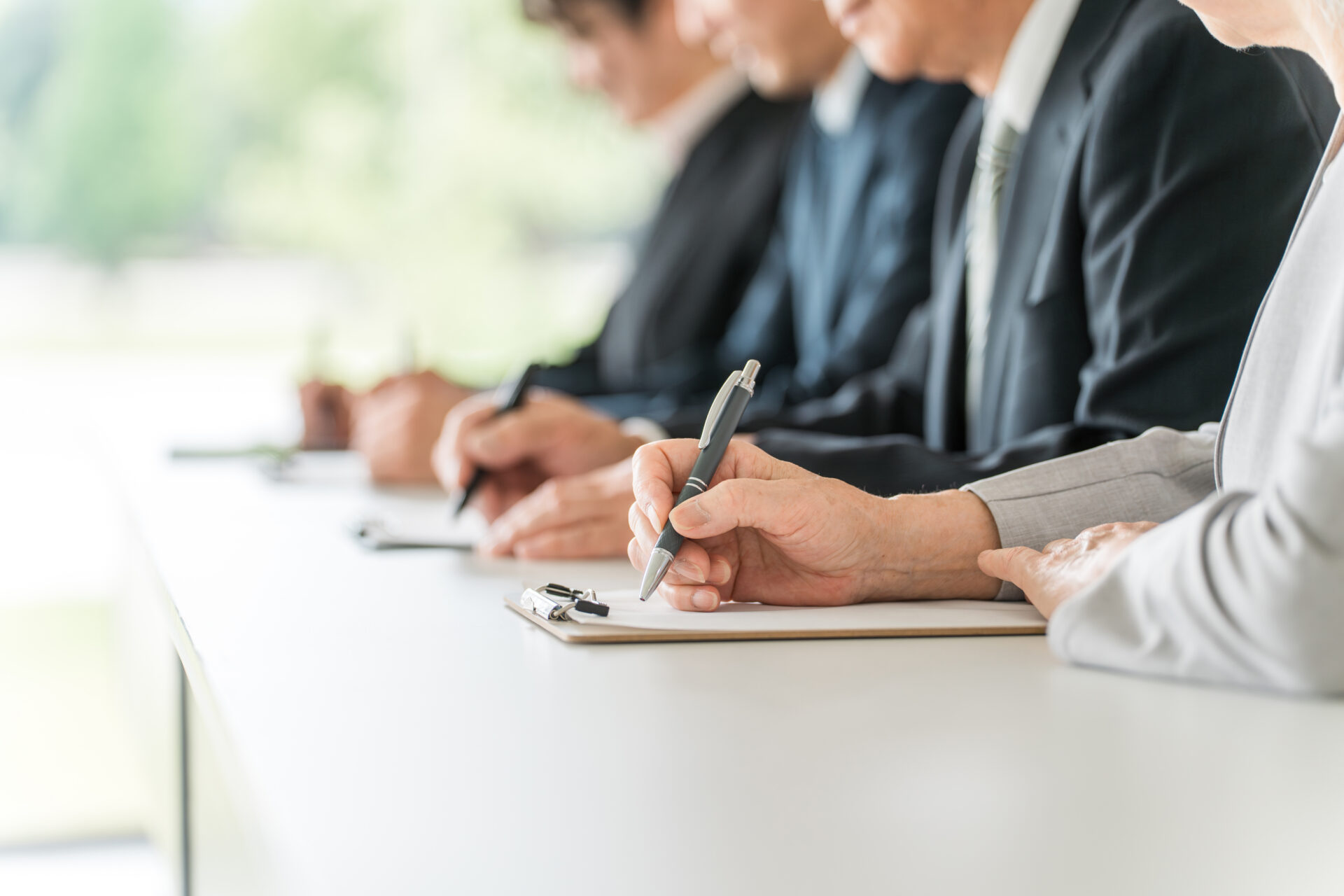 Group of professionals in business attire seated at a table, taking notes with pens on clipboards.