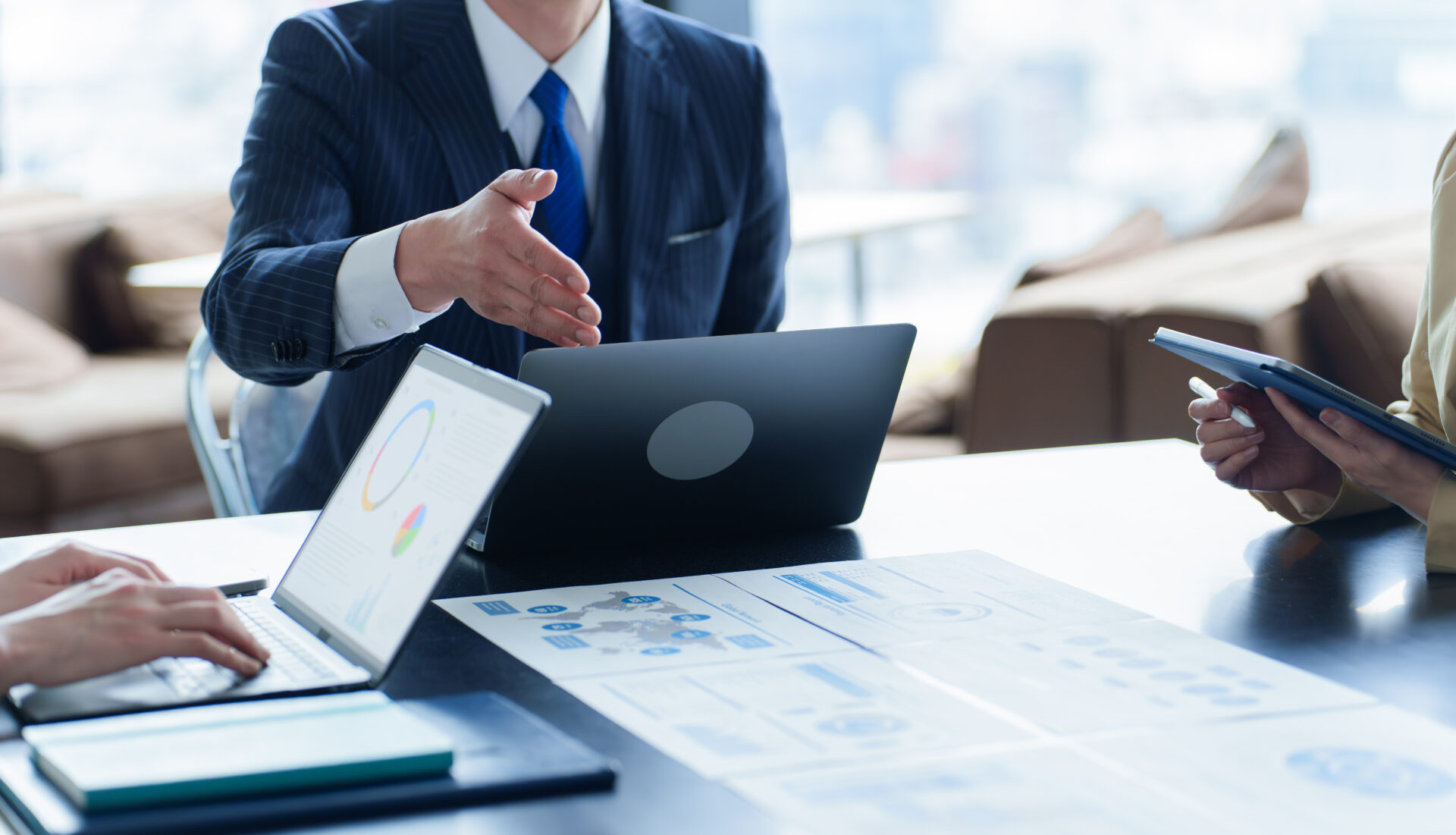 Business meeting scene with a man in a pinstripe suit extending a handshake over a table with laptops and printed charts.