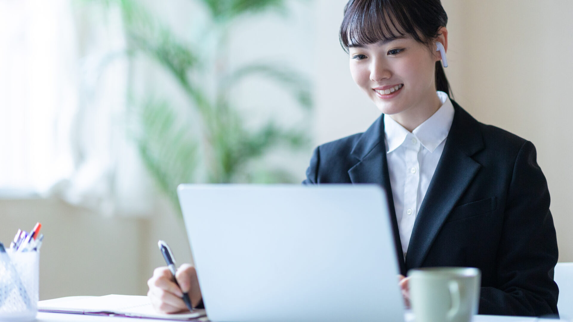 仕事をする女性 | 日本最初の品質マネジメントシステム認証機関 -日本化学キューエイ株式会社 Woman in a business suit using a laptop at a desk, wearing a headset and writing in a notebook.