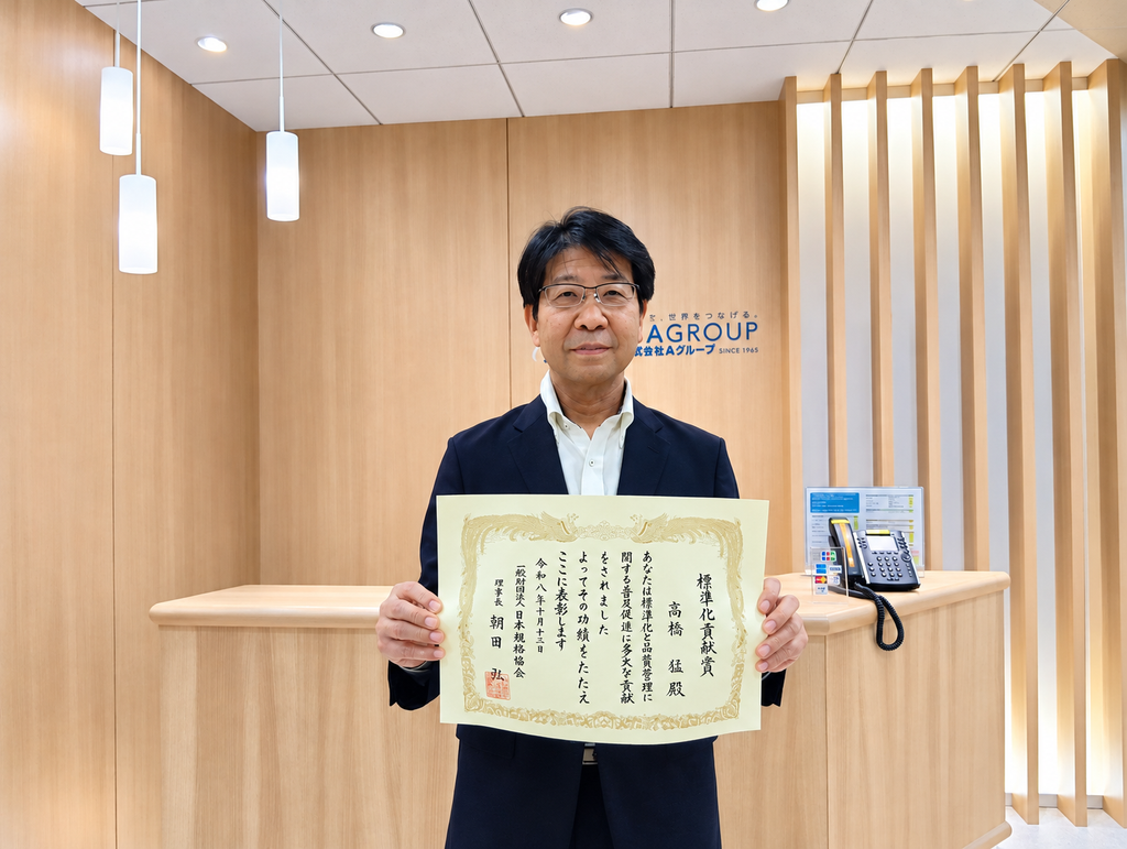 高橋顧問画像賞状 | 日本最初の品質マネジメントシステム認証機関 -日本化学キューエイ株式会社 Man in a dark suit holds a large ceremonial certificate in a wood-paneled reception area with a blue AGROUP sign behind him.